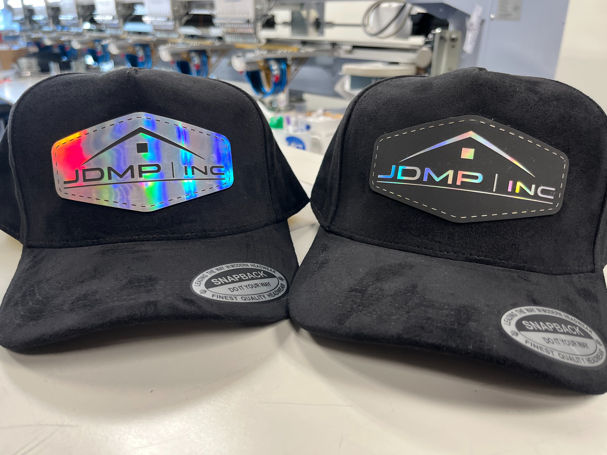 Two all-black suede hats with custom laser-engraved company holographic logo patches