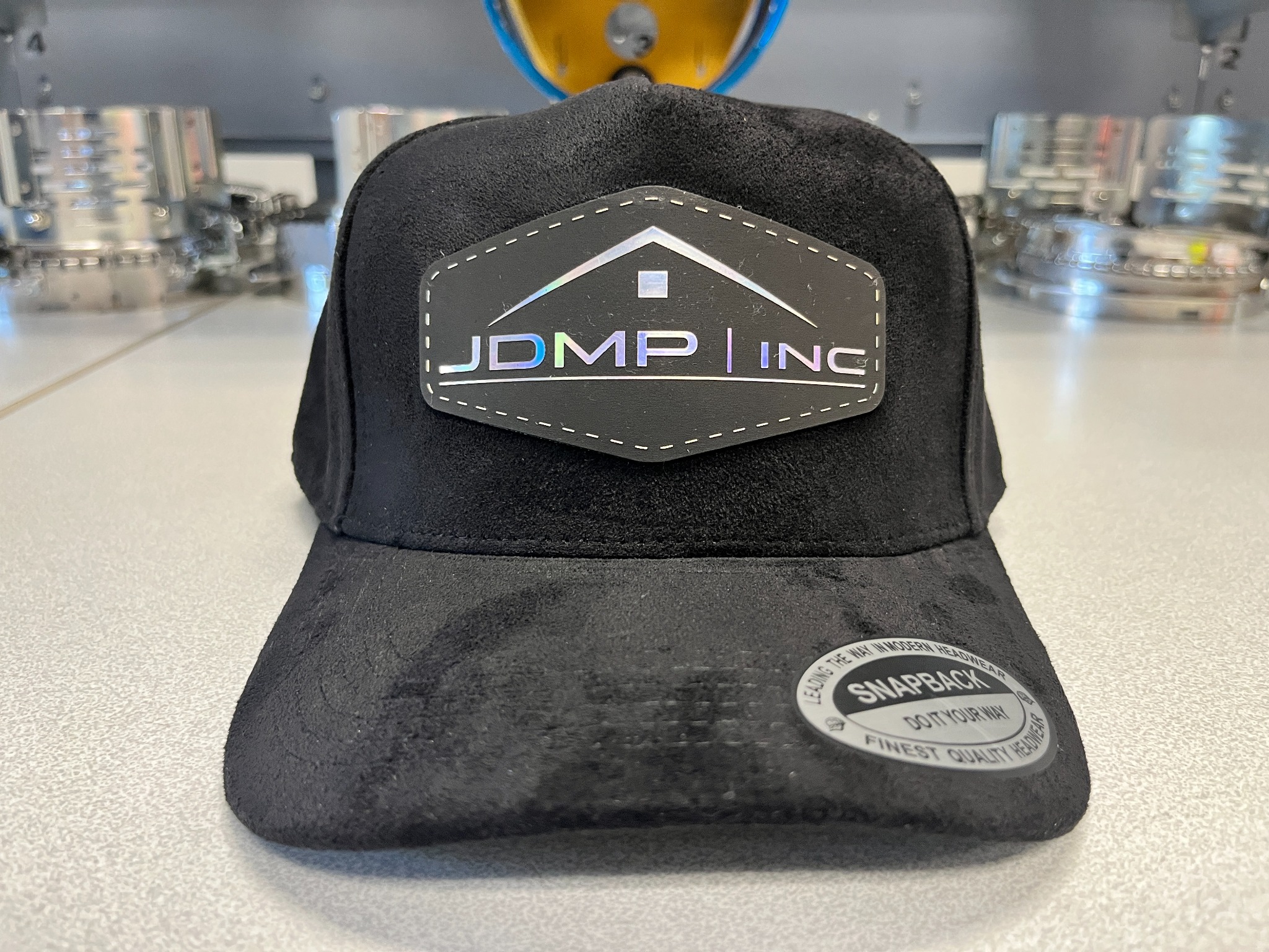 Black suede hat with a shaped dark company patch and iridescent logo detail