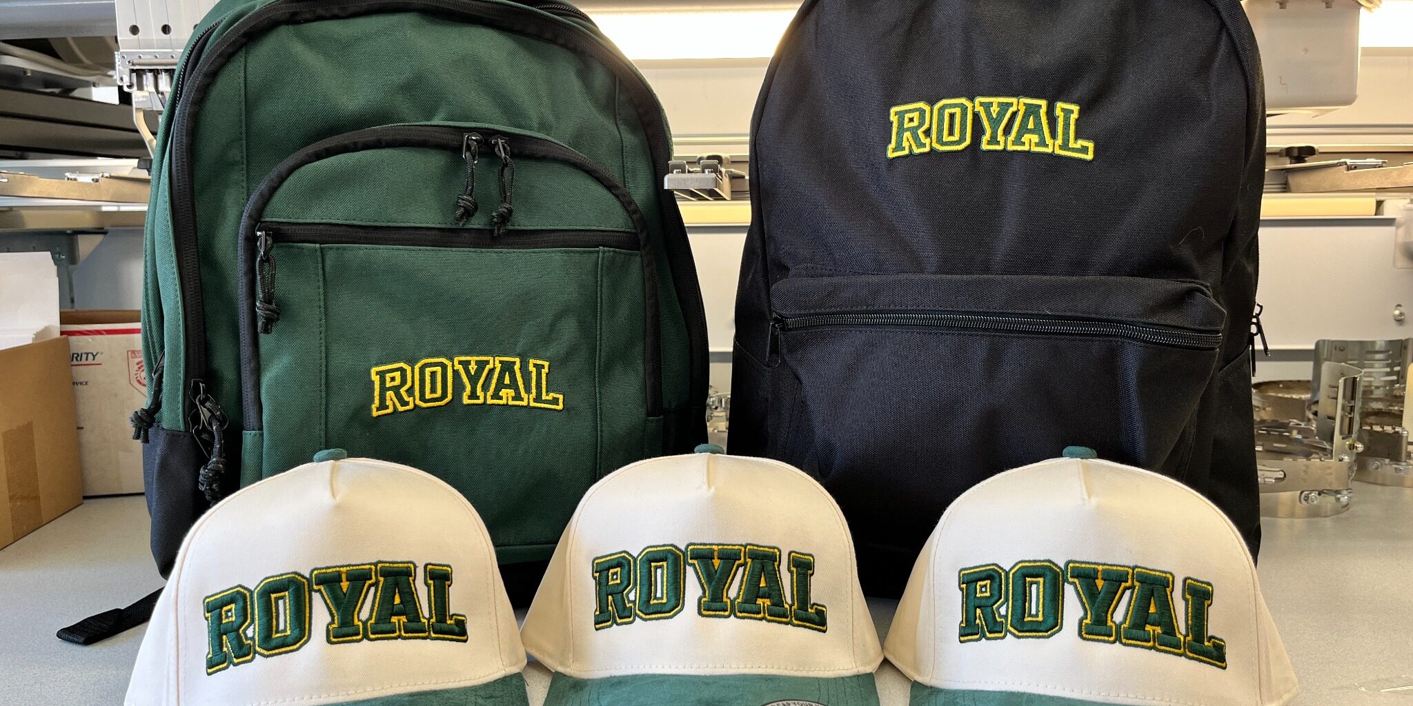 Green and black embroidered Royal High School backpacks behind cream and green Royal hats