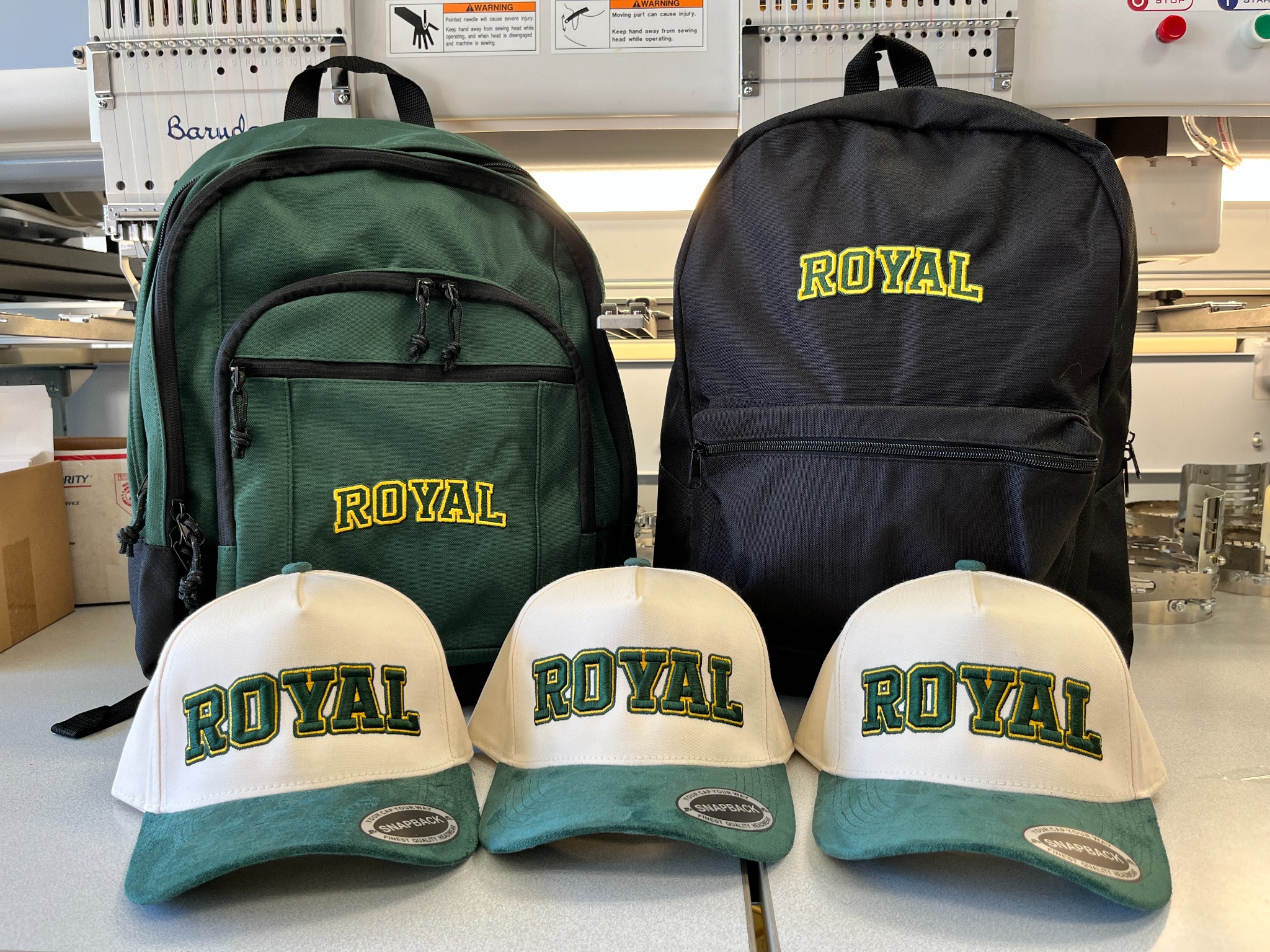 Green and black embroidered Royal High School backpacks behind cream and green Royal hats