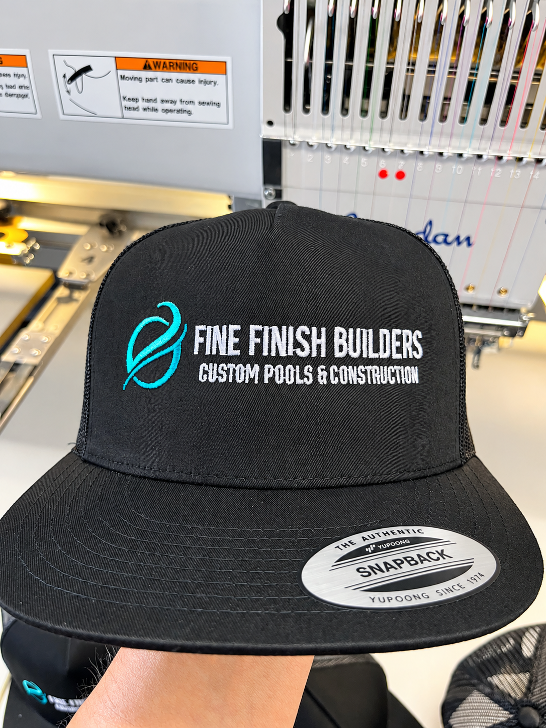 Black snapback hat with turquoise and white Fine Finish Builders embroidery in front of a Barudan machine.