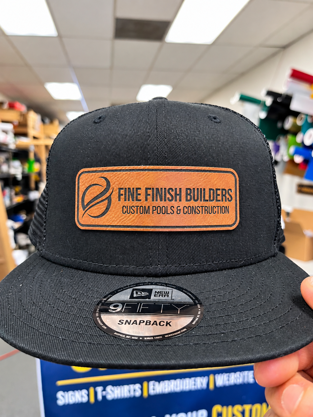 Black New Era cap with a brown Fine Finish Builders logo patch by SpectraColor.